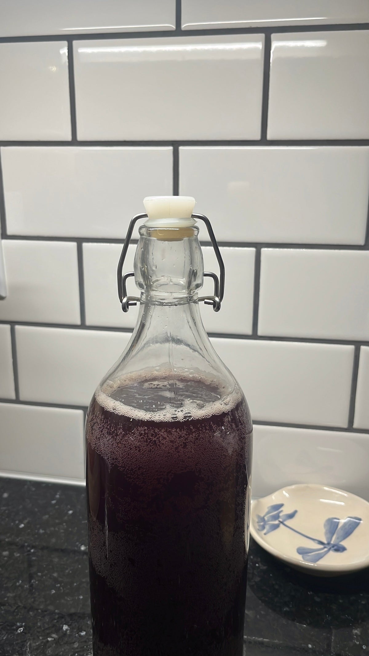 Purple fermented cherry soda carbonating in a clear swing-top bottle against white tile.